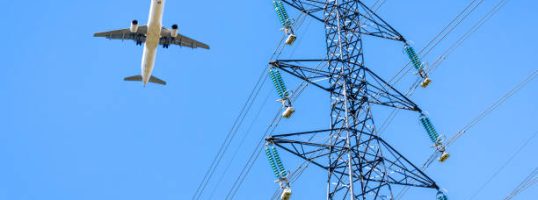 View from below of an airliner in landing approach flying over a high-voltage power line against blue sky with an electricity pylon in the foreground.